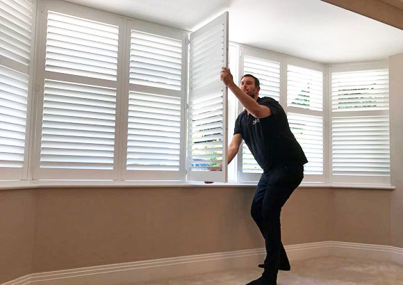 A soft light fills the room as a man stands close to a shuttered window, gazing out.