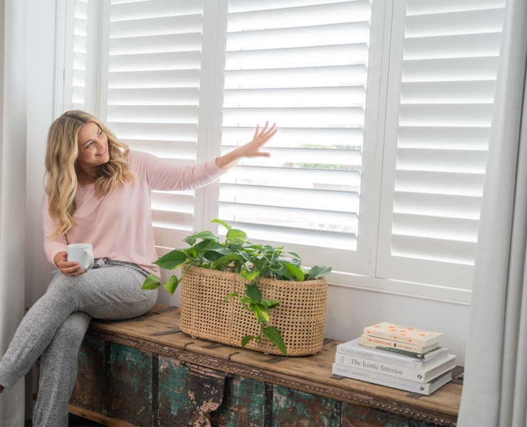 A serene moment is captured indoors as a woman sits on a bench and looks out through a shuttered window.